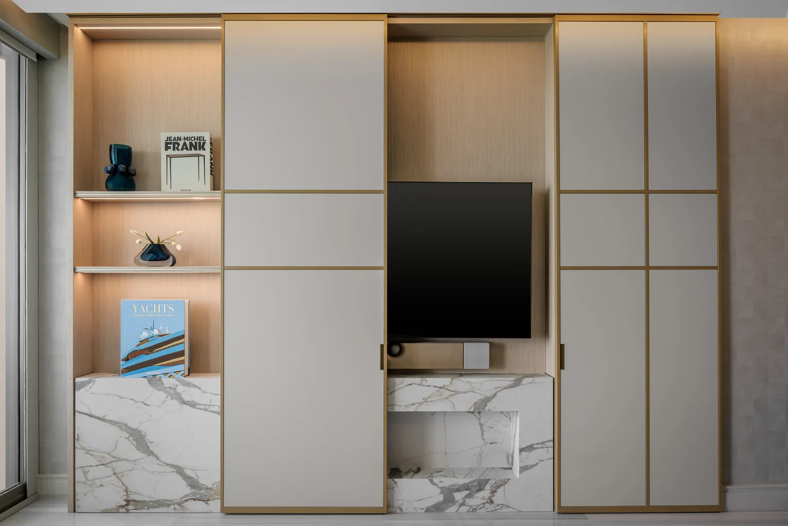 Living room millwork with brass-framed sliding panels open to a TV niche, lit oak shelves and marble base, Acqualina 2303, Sunny Isles Beach